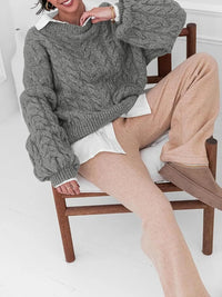 Woman in a cozy cable knit long sleeve sweater paired with soft beige pants, sitting on a wooden chair.