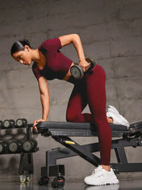 Woman exercising in a burgundy round neck short sleeve top and leggings set with dumbbell on a workout bench.