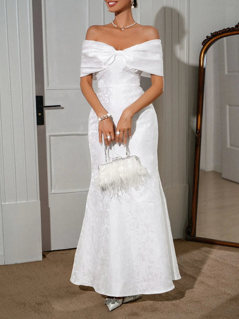 Elegant woman wearing a white bow off-shoulder maxi dress, holding a stylish handbag, in a chic indoor setting.