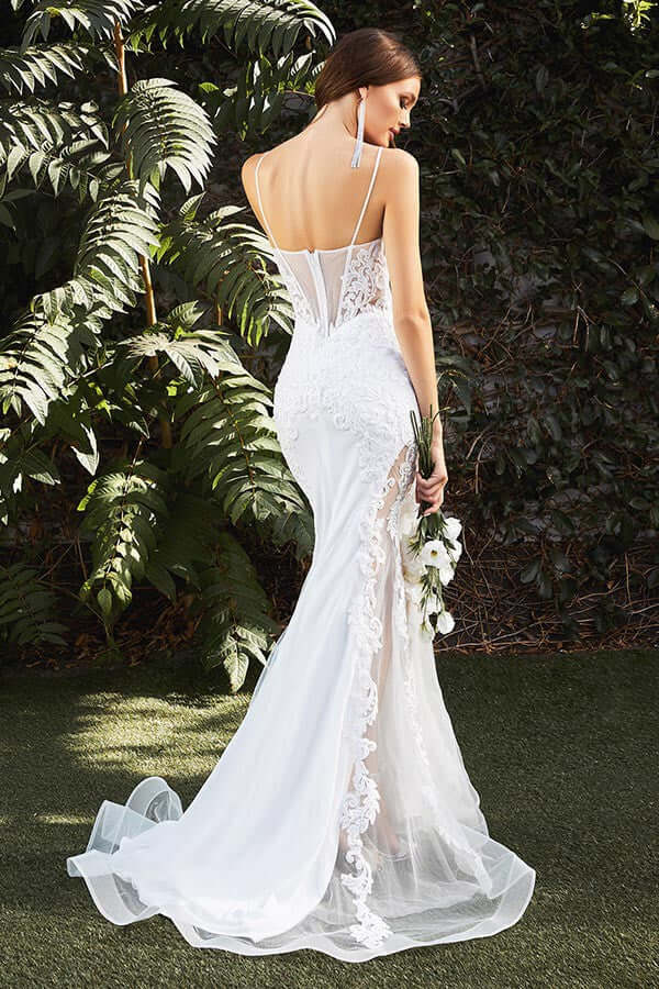 Woman in a white lace-back mermaid wedding gown/dress holding flowers, standing outdoors in a lush green garden.