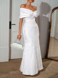 Elegant off-shoulder maxi dress in white with bow detail, showcased in a stylish interior.
