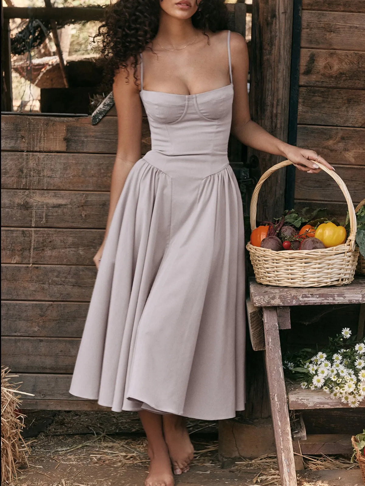 Model wearing a ruched sweetheart neck cami dress in light taupe, holding a basket of vegetables in a rustic setting.