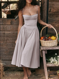 Model wearing a ruched sweetheart neck cami dress in light taupe, holding a basket of vegetables in a rustic setting.