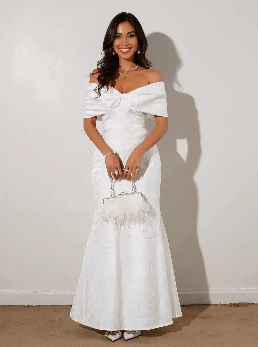 Model wearing a white off-shoulder maxi dress with a bow, holding a handbag and smiling.