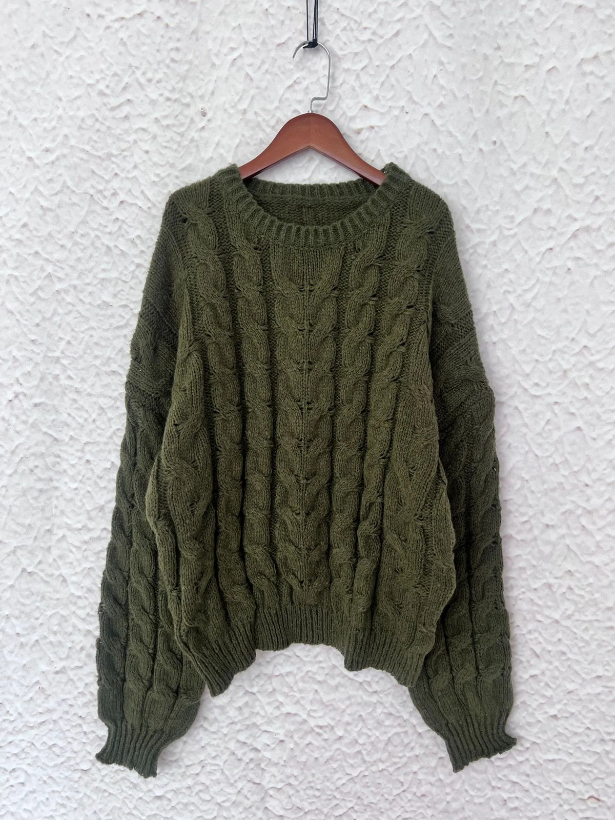Olive green cable knit long sleeve sweater displayed on a wooden hanger against a textured white background.