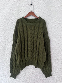 Olive green cable knit long sleeve sweater displayed on a wooden hanger against a textured white background.