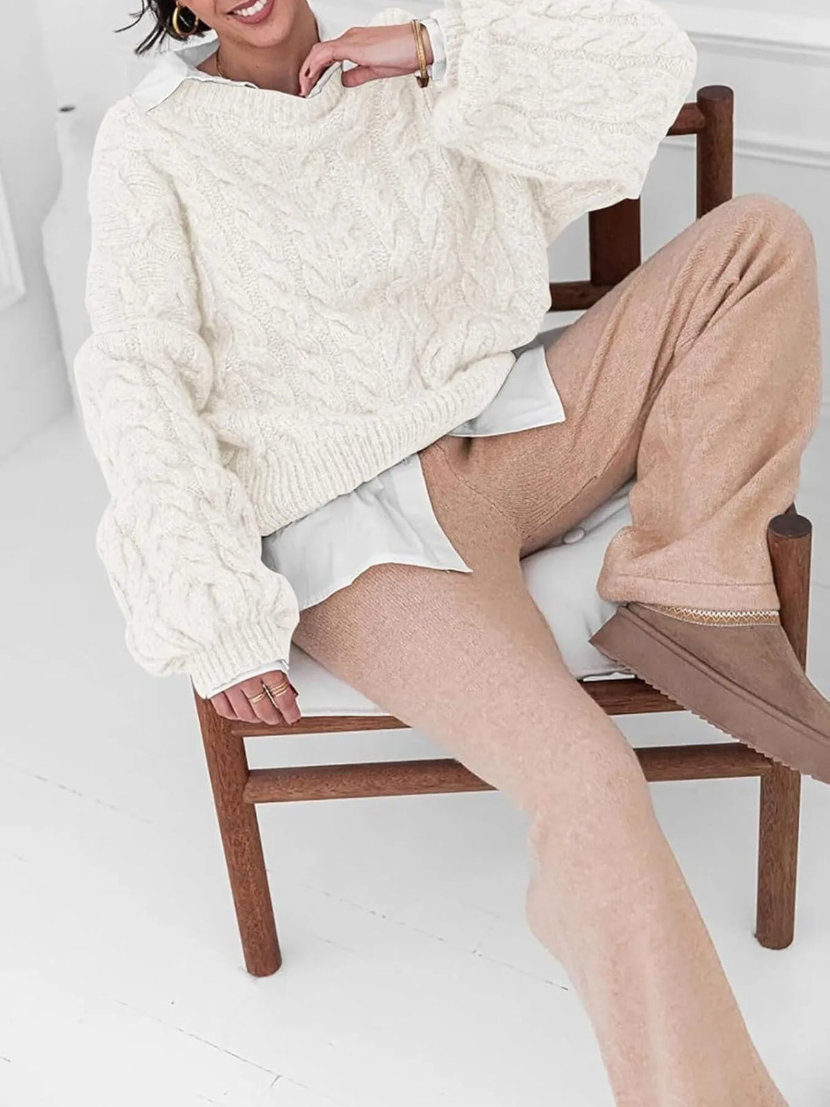 Model wearing a cream cable knit long sleeve sweater paired with beige pants, sitting on a wooden chair.