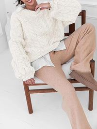 Model wearing a cream cable knit long sleeve sweater paired with beige pants, sitting on a wooden chair.