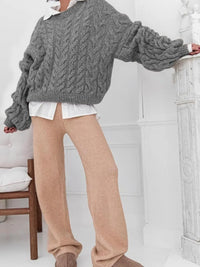 Model wearing a cable knit long sleeve sweater with oversized sleeves and beige pants in a stylish indoor setting.