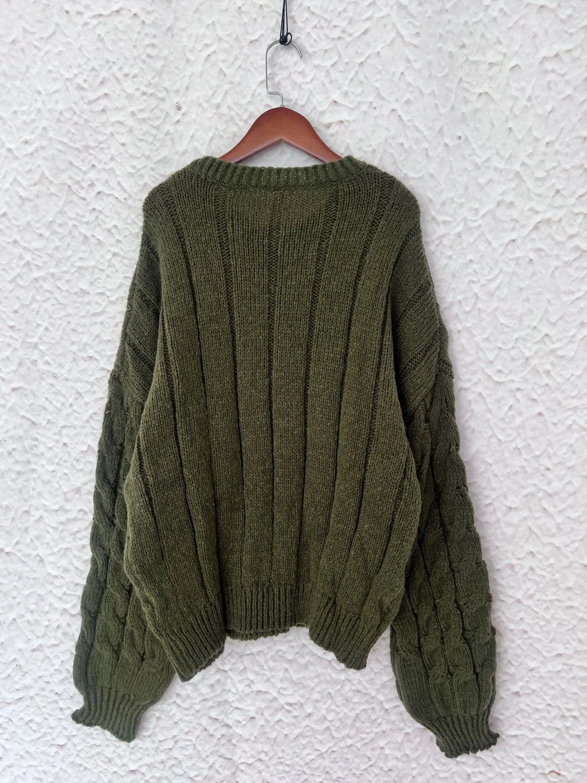 Back view of a green cable knit long sleeve sweater hanging on a hanger against a textured wall.