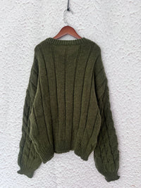 Back view of a green cable knit long sleeve sweater hanging on a hanger against a textured wall.
