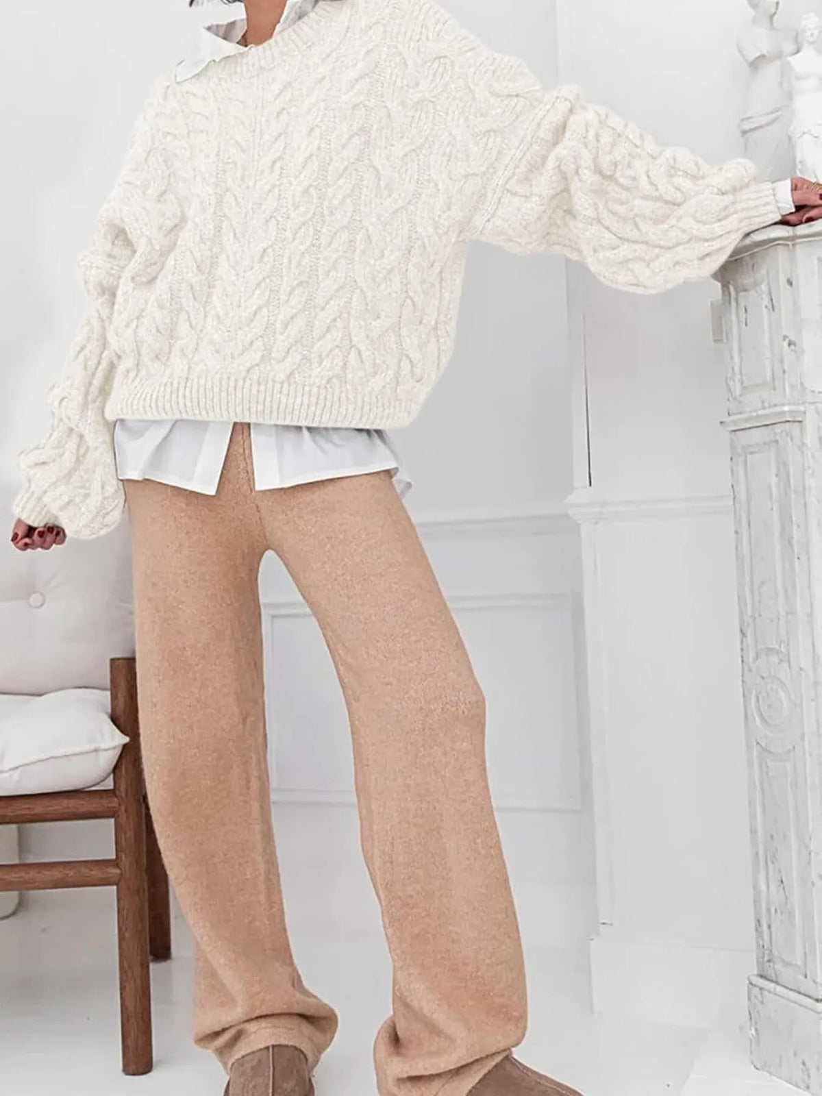 Model wearing a cream cable knit long sleeve sweater paired with beige wide-leg pants in a bright, minimalist setting.