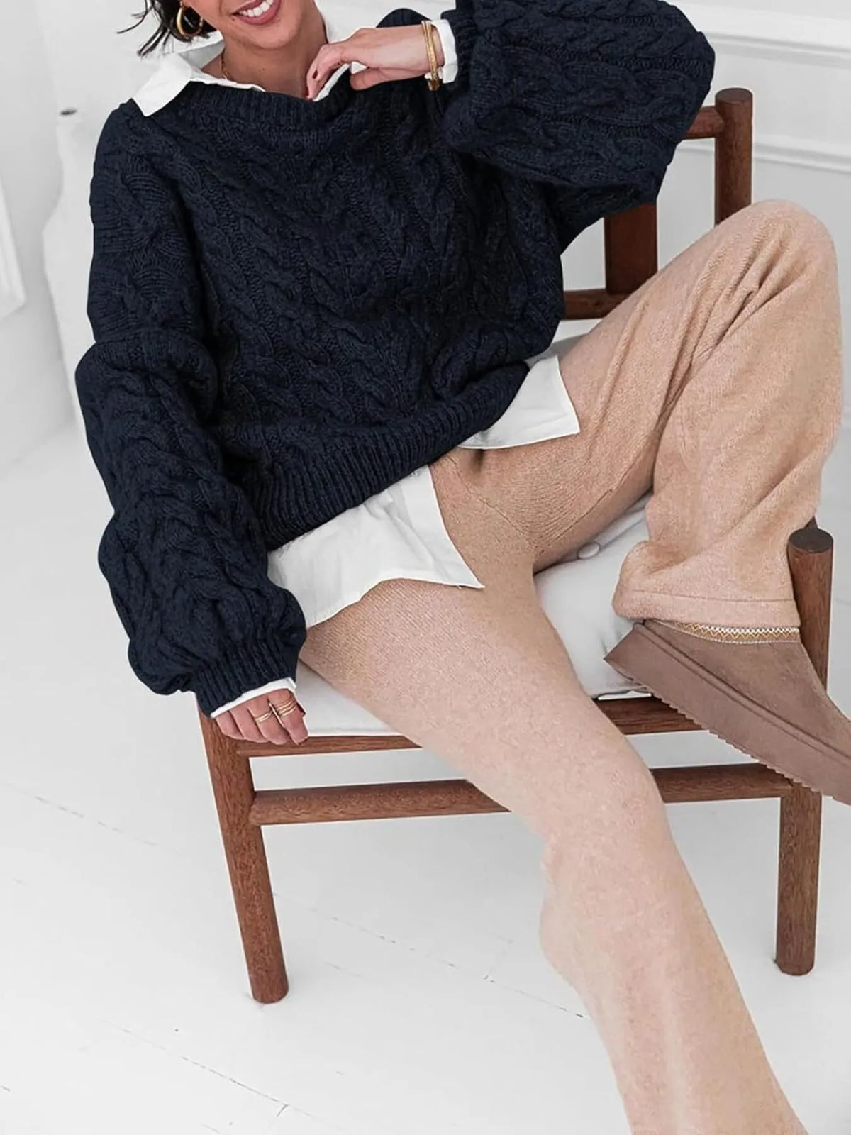 Model wearing a navy cable knit long sleeve sweater paired with beige pants, seated on a wooden chair.