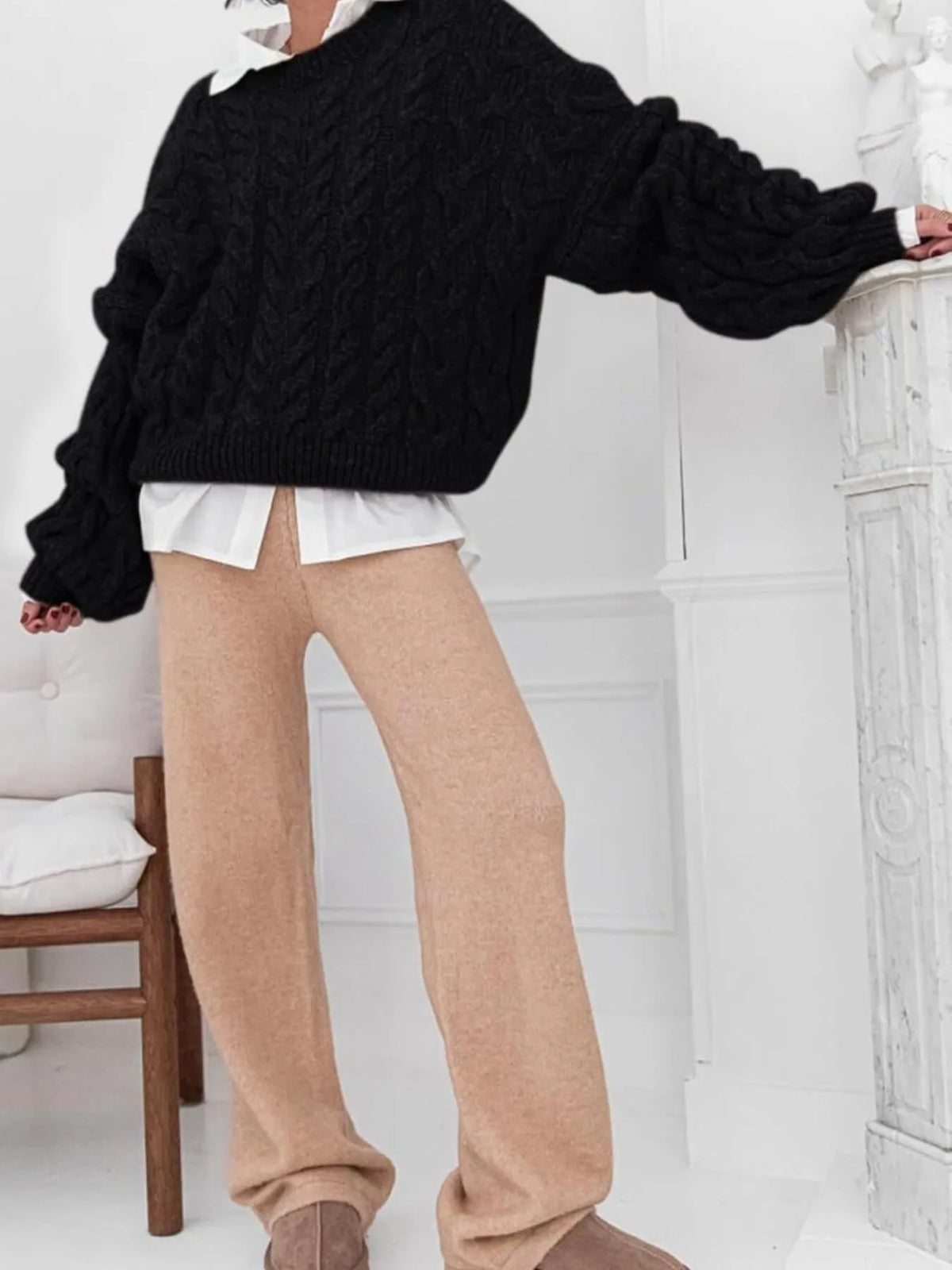 Model wearing a black cable knit long sleeve sweater paired with beige pants in a bright, minimalistic setting.