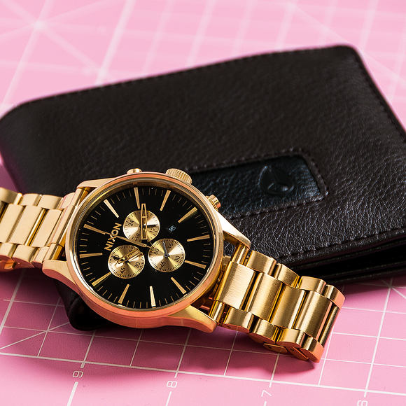 Nixon Sentry Chrono All Gold & Black Men's Watch A386-510 on a pink surface beside a black wallet.