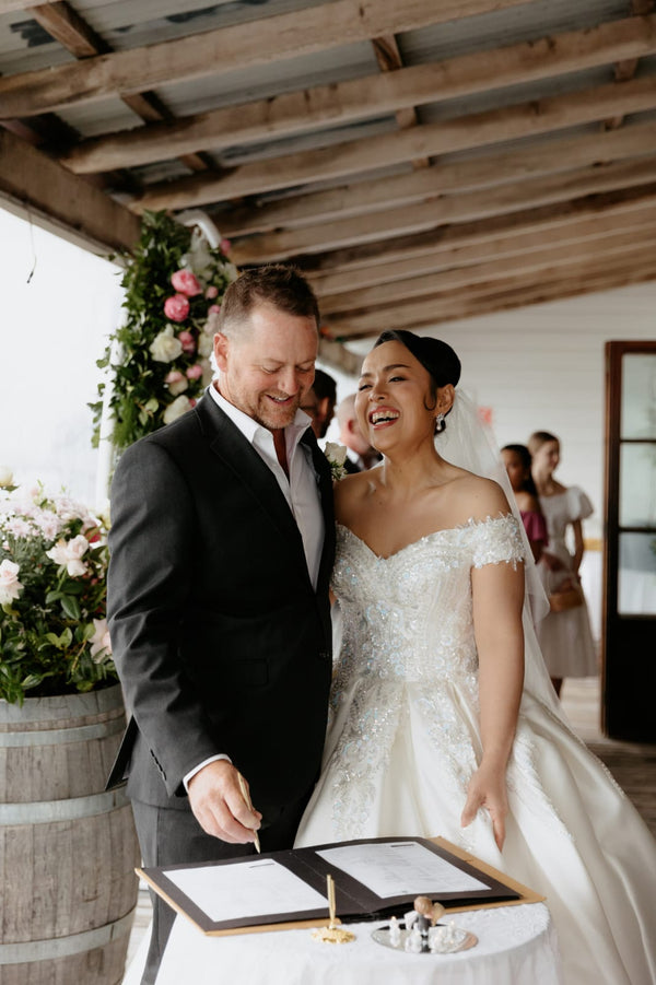 Couple signing a document at a wedding ceremony with floral decorations and guests in the background.